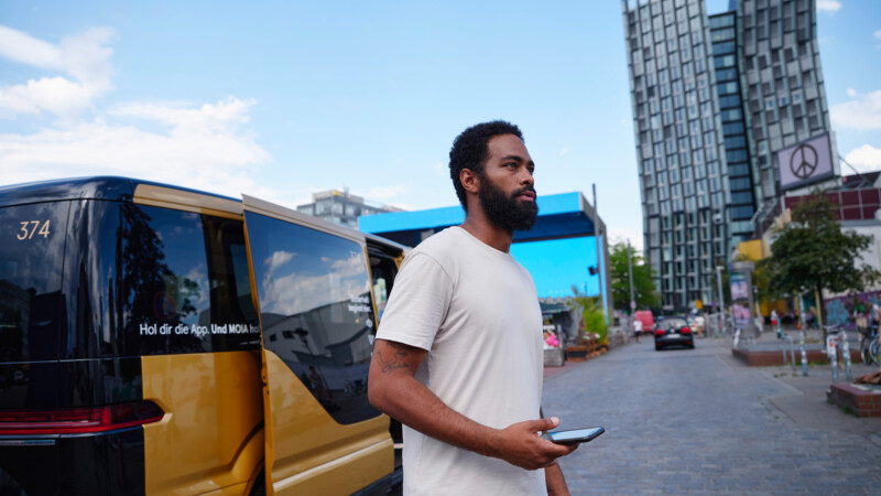 Schwarzer Mann steht vor Moia-Taxi mit Handy in der Hand und den Tanzenden Türmen im Hintergrund